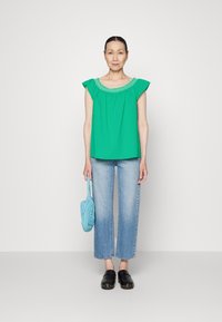 Blusa verde com um decote texturizado, combinada com jeans lisos de perna reta azuis claros e tamancos pretos. A segurar uma mala de rede azul clara.