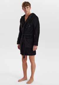 Black cotton bathrobe with a hood, belted waist, front pockets, and subtle branding on the chest. Soft texture and mid-thigh length.