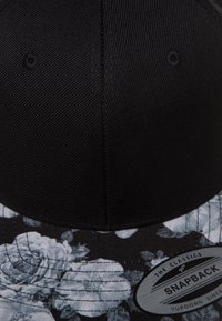 Schwarze Snapback-Cap mit einem floralen Schwarz-Weiß-Muster am Schirm, einem strukturierten Stofffinish und einem Markenetikett an der Seite.