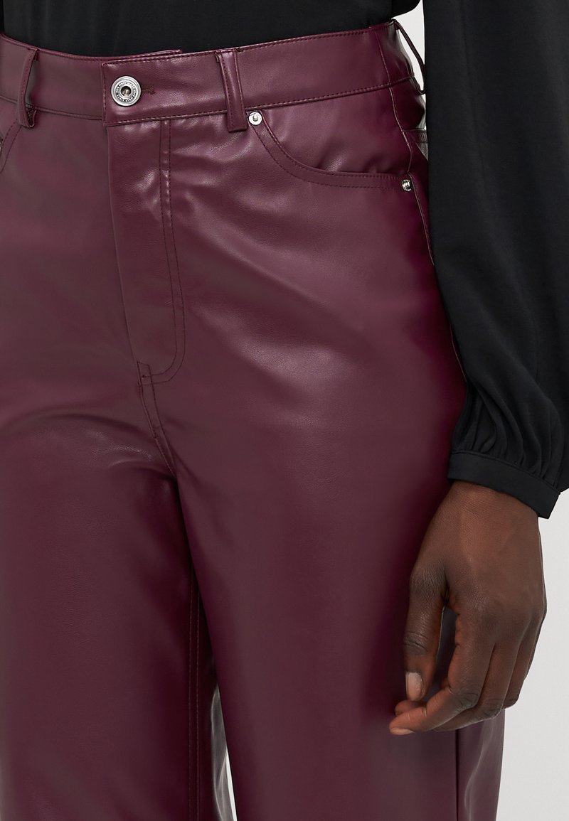 Pantaloni in faux leather borgogna con design aderente, tasche frontali e chiusura con bottone. La superficie ha una texture liscia e lucida.