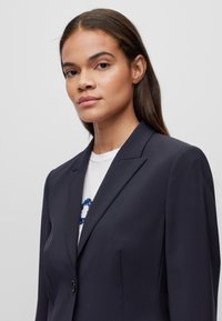 Blazer navy con rever a punta, chiusura con un bottone e spalle strutturate, realizzato in tessuto liscio e con una vestibilità sartoriale sopra una camicia bianca.