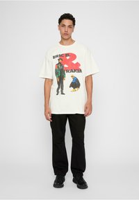 Lys beige grafisk t-shirt med et levende print af to figurer og tekst, parret med sorte cargo bukser og sorte sneakers. Løs pasform.