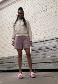 Lys beige zip-up hættejakke, strukturerede lyserøde shorts og lyserøde platform sandaler. Sat foran en hvid murstensvæg og metalstillads.