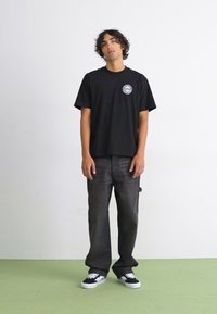 Schwarzes T-Shirt mit kurzen Ärmeln und einem kreisförmigen Grafikdesign, kombiniert mit grauen, locker sitzenden Jeans und schwarzen Sneakers mit weißen Sohlen.