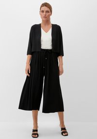 Zwarte verkorte cardigan over een witte V-hals top, gecombineerd met zwarte plooibroek met wijde pijpen. De stijl bevat een elastische taille en platte sandalen.