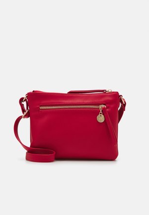 Rød læder crossbody-taske med gyldent lynlås og cirkulært vedhæng på frontlommen, justerbar rem og glat finish på ensfarvet baggrund.