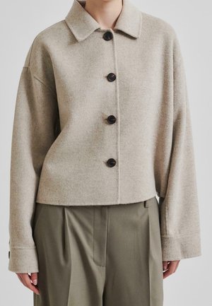 Beige uld kort jakke med fire sorte knapper, bred krave og lange ærmer, båret med olivengrønne plisserede bukser.