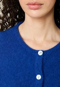 Blauwe, gestructureerde cardigan met een ronde hals en twee witte knopen aan de voorkant. De stof lijkt zacht en comfortabel.