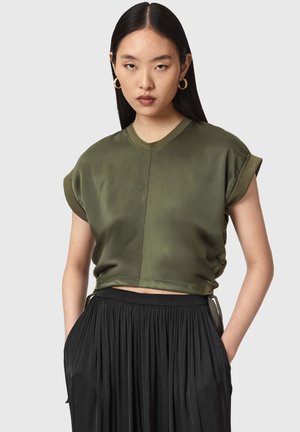 Jeune femme aux cheveux noirs raides porte un haut sans manches vert olive et une jupe plissée noire, debout les mains dans les poches.