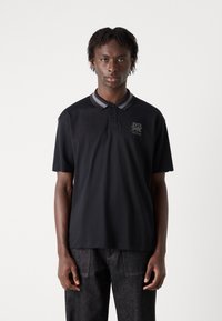 Zwarte polo met een bidimensionale kraag, voorzien van een subtiele opdruk op de borst. Het materiaal lijkt glad en heeft korte mouwen.