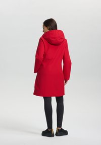 Manteau imperméable rouge avec capuche, dotée d'un col texturé, de manches longues et de poches latérales, associé à un legging noir et des sneakers.