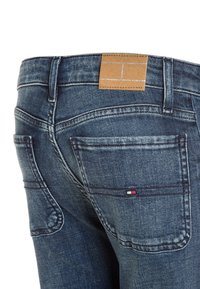 Jeans di denim con lavaggio blu scuro, caratterizzati da una toppa in pelle marrone in vita e due tasche posteriori con dettagli di cucitura.
