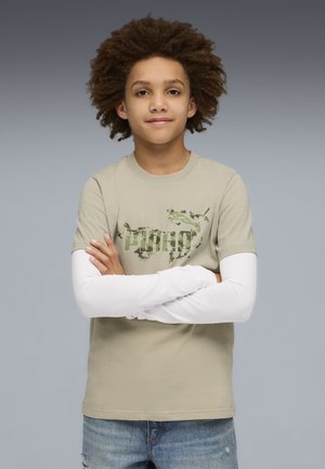 Chłopiec z kręconymi włosami, ubrany w beżową koszulkę Puma na białej, długim rękawie i niebieskich dżinsach, z założonymi rękami na szarym tle.