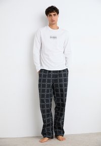 Cămașă albă cu mâneci lungi, având un logo negru pe piept, asortată cu pantaloni de pijama cu carouri negre, având linii albe în grilă.