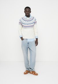 Μπεζ πλεκτό πουλόβερ με γεωμετρικό μοτίβο σε μπλε και ροζ, φορεμένο πάνω από ανοιχτό μπλε πουκάμισο με γιακά, συνδυασμένο με ανοιχτό μπλε τζιν και καφέ παπούτσια.