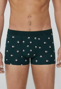 Männlicher Oberkörper trägt dunkelgrüne Boxershorts mit einem Muster aus verstreuten schwarz-weißen Billardkugeln mit der Nummer 8.