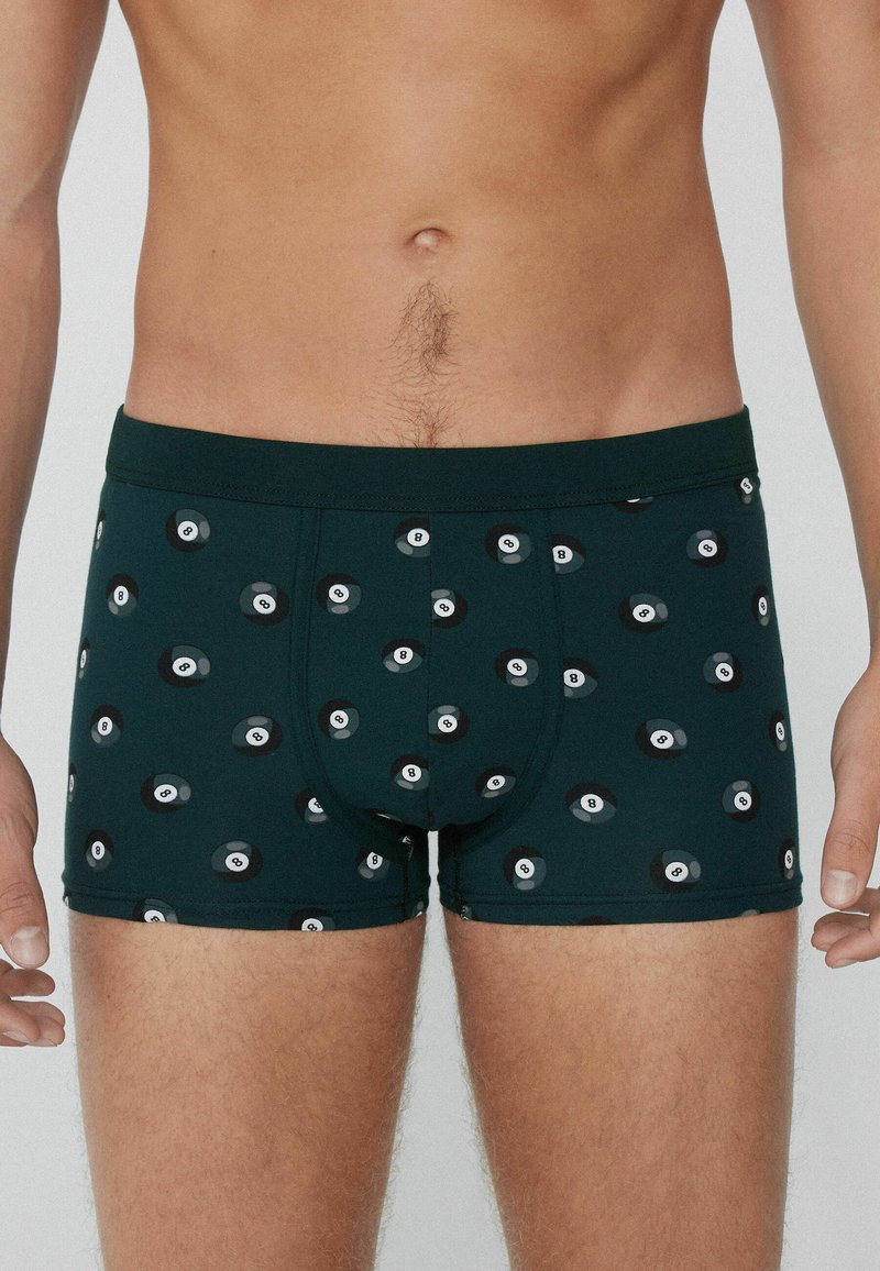 Männlicher Oberkörper trägt dunkelgrüne Boxershorts mit einem Muster aus verstreuten schwarz-weißen Billardkugeln mit der Nummer 8.
