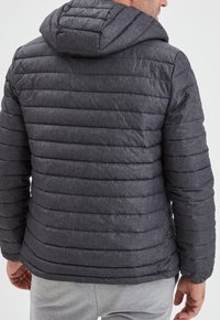 Giacca a vento grigia con pannelli a cucitura orizzontale e cappuccio. Il materiale ha una finitura testurizzata. Polsini e orlo aperti con un design moderno.