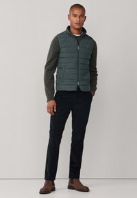 Donkergroene gewatteerde bodywarmer over een grijze trui, gecombineerd met marineblauwe broek en bruine Chelsea laarzen. Voorzien van een ritssluiting en zijzakken.