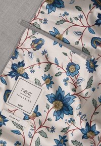 Tessuto floreale con fiori blu e crema e foglie verdi, bordato con una rifinitura grigio chiaro. Etichetta recante la scritta "next tailoring, 40R, slim fit."