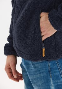 Laivasto sininen fleece-puluveri, jossa on tekstuuripinta, varustettuna vetoketjullisella taskulla ja vaaleanruskealla vetonauhalla, yhdistettynä vaaleansinisiin farkkuihin.