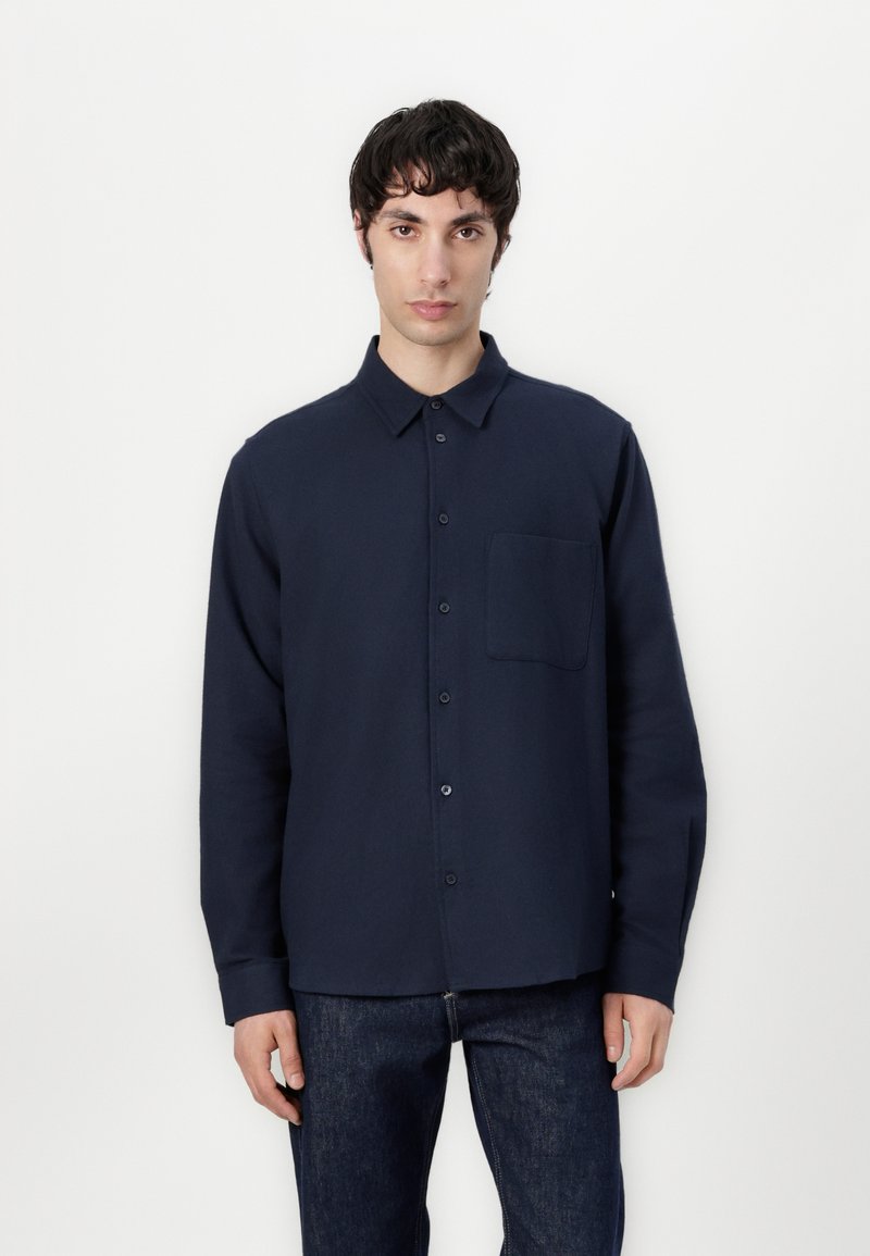 Navy long-sleeve shirt with button-up closure, collar, single chest pocket, and a smooth texture. Worn with dark denim jeans.