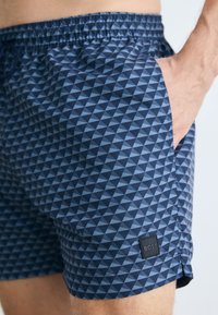 Tengeri kék fürdőnadrág, amely geometrikus háromszög mintával rendelkezik, különböző kék árnyalatokban. Gumis derékpánt állítható zsinórral és oldalsó zsebekkel.
