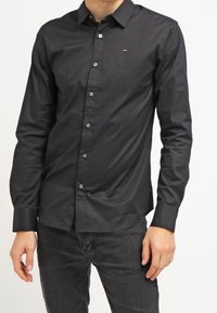 Camisa negra de manga larga con botones y corte entallado, que presenta un logo sutil en el pecho. Textura suave, cuello clásico y puños estándar.