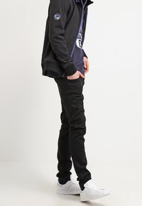 Svarta jeans, svart zip-up jacka med blå accenter, vita sneakers och en marinblå skjorta. Tätt sittande design med minimala detaljer.