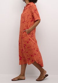 Korte mouwen, gekleed jurk met kraag in feloranje met een bladdessin, voorzien van zijzakken en een gebogen zoom. Draagt met bruine sandalen.