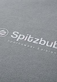 Tessuto grigio con un motivo a maglia testurizzata caratterizzato da un logo ricamato bianco "Spitzbub Sportswear Edition" e un emblema circolare.