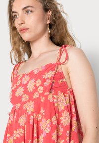 Vestido floral rosa com alças amarradas, apresentando um corpete franzido e uma bainha plisada fluida. Padrão floral em amarelo e rosa.