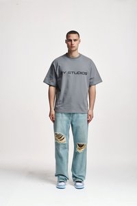 Lühikeste juustega mees seisab näoga otse, seljas hall "2Y STUDIOS" t-särk, heledast sinisest rebenenud teksapüksid ja siniste ja valgete jooksujalatsid.