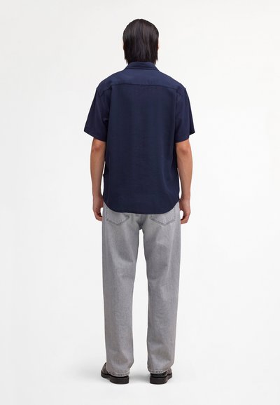 Man standing facing away wearing a navy short-sleeve shirt, light gray jeans, and black shoes on a plain white background.