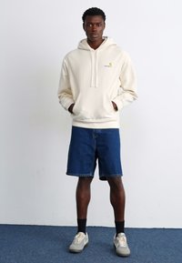 Lys beige hættetrøje med lomme foran, parret med mørkeblå denimshorts. Modellen har grå sneakers og sorte sokker på.
