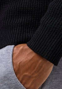 Schwarzer, gerippter Pulloverwulst, der über eine graue Stofftasche überlappt, mit einer sichtbaren Hand, die gegen den Stoff ruht und Hautstruktur zeigt.