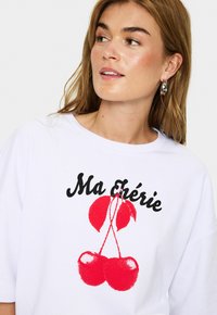 Baltas kokvilnas T-krekls ar melnu skriptu "Ma Chérie" un sarkanu ķiršu grafiku ar teksturētu dizainu. Brīvs piegriezums ar apaļu dekoltē.