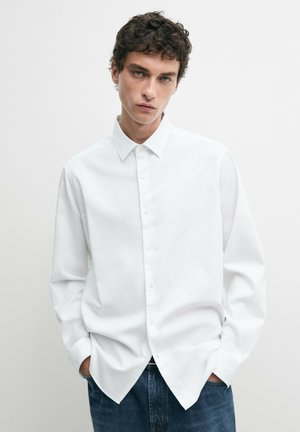 Giovane uomo con capelli ricci che indossa una camicia bianca semplice con bottoni e jeans blu, in piedi con le mani in tasca contro uno sfondo bianco.
