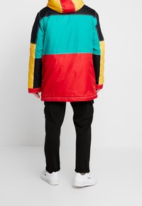 Färgglad parka-jacka med sektioner i röd, grön, gul och svart. Slät tyg, huva och kontrasterande sömmar. Kommer ihop med svarta byxor och vita sneakers.