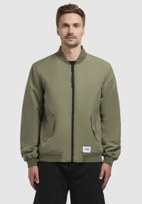 Olivgrüne Bomberjacke mit Reißverschluss vorne, geripptem Kragen und zwei Seitentaschen. Mit elastischen Bündchen und einem Logosticker am Saum.