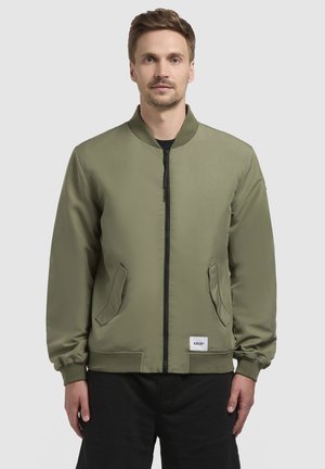 Maslinasto zelena bomber jakna s prednjim patentnim zatvaračem, rebrastim ovratnikom i dva bočna džepa. Sadrži elastične manžete i oznaku s logotipom na rubu.