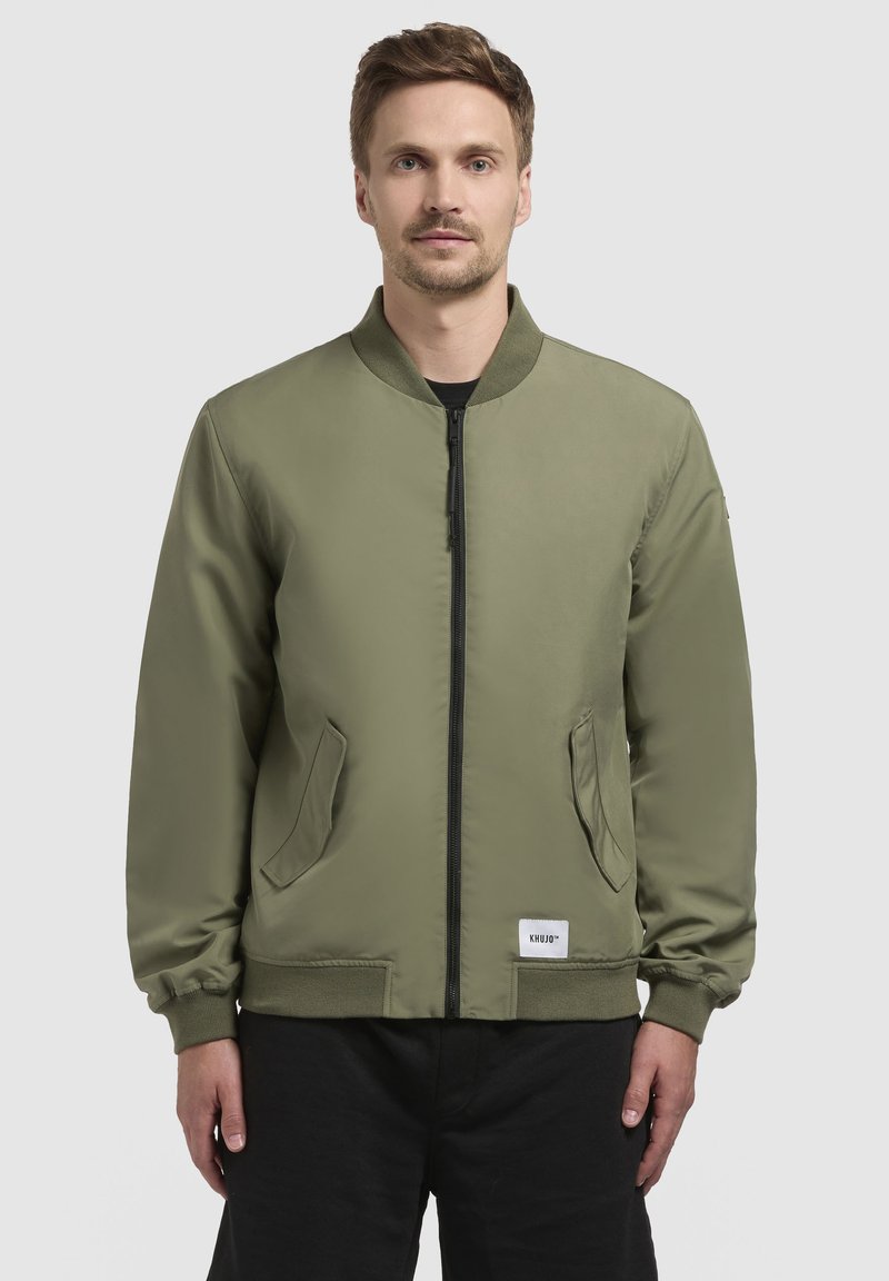 Olivovozelená bomber bunda so zipsom vpredu, prúžkovaným golierom a dvoma bočnými vreckami. Má elastické manžety a štítok s logom na leme.