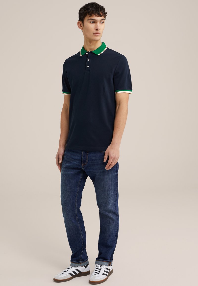 Homme debout portant un polo marine avec un col et des bordures de manches vertes, un jean bleu foncé et des baskets blanches avec des rayures noires.