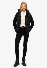 Zwarte pufferjack met gestructureerde mouwen, ritsluiting en capuchon, gedragen over een crèmekleurige kabelgebreide trui en zwarte getailleerde broek met laarzen.