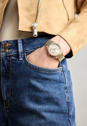 Montre-bracelet analogique en ton or avec un cadran rond, trois sous-cadrans et un bracelet en métal texturé. Portée avec une veste en daim beige et un jean en denim bleu.