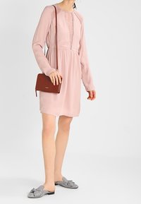 Rosa långärmad klänning med texturerade detaljer, åtstramad midja och lätt material; matchad med grå mules med rutmönster och en brun crossbody-väska.