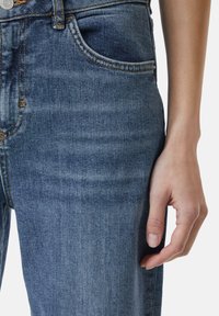 Dunkelblaue Denim-Jeans mit glatter Textur, ausgestattet mit einer Vordertasche und Messingbeschlägen an der Taille. Hand ruht an der Seite.