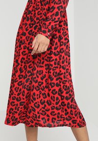 Robe midi rouge avec des taches de léopard noires irrégulières, manches longues et poignets boutonnés, portée par une personne avec une main et des jambes visibles.