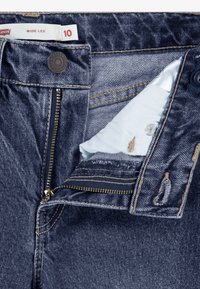 Mörkblå denimjeans med en öppen mässingsdragkedja, delvis öppnad tygklaff och etikett som visar "Wide Leg" och storlek 10.