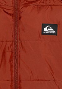 Rød puffjakke med en jevn, shiny overflate. Har en sentral glidelås og en svart Quiksilver-logo på venstre side.